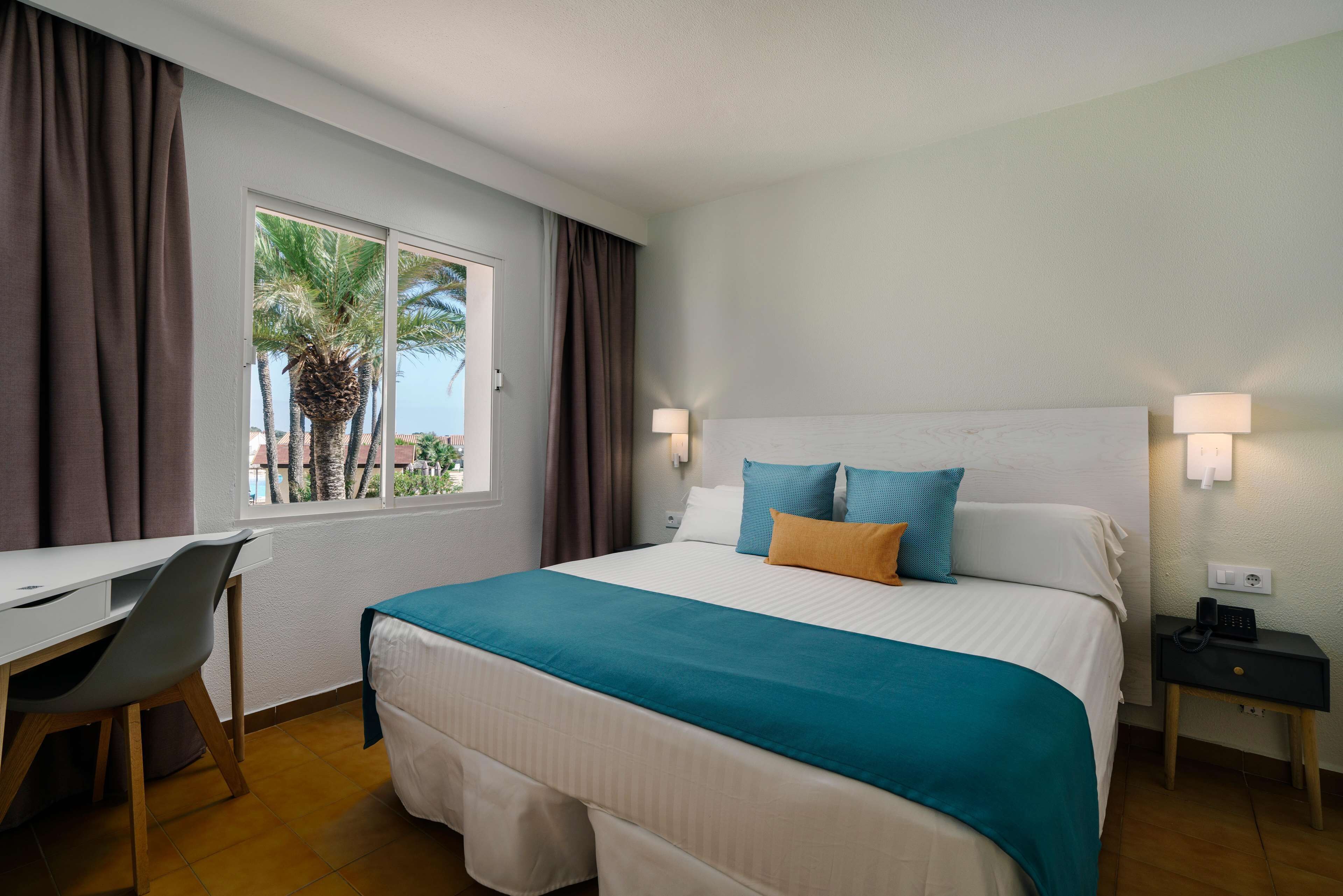 Una habitación de hotel con cama, escritorio, silla, mesita de noche y ventana con cortinas.