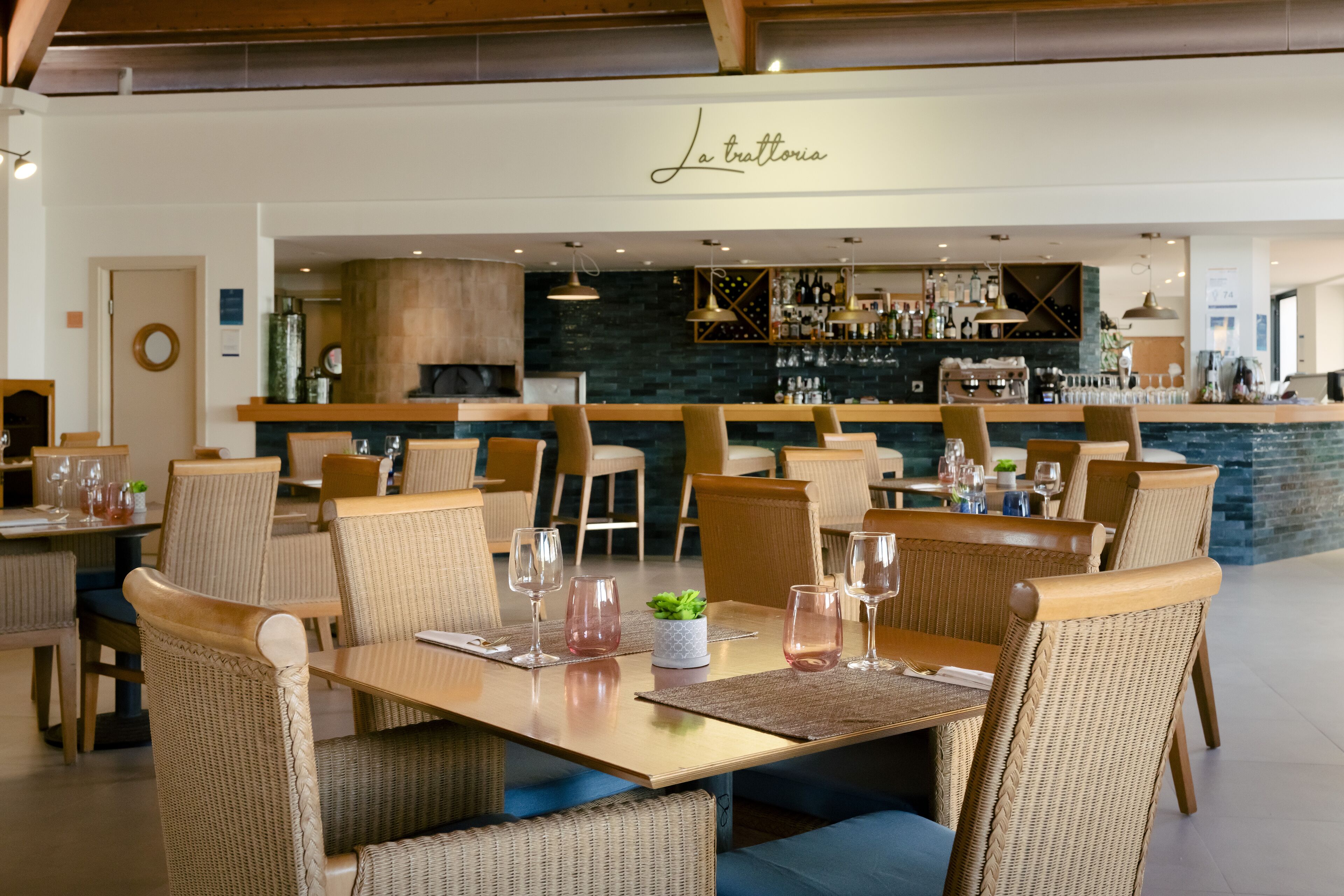 Un restaurante con sillas y mesas de mimbre, una zona de bar con botellas y vasos, y un letrero que dice 'La trattoria'.