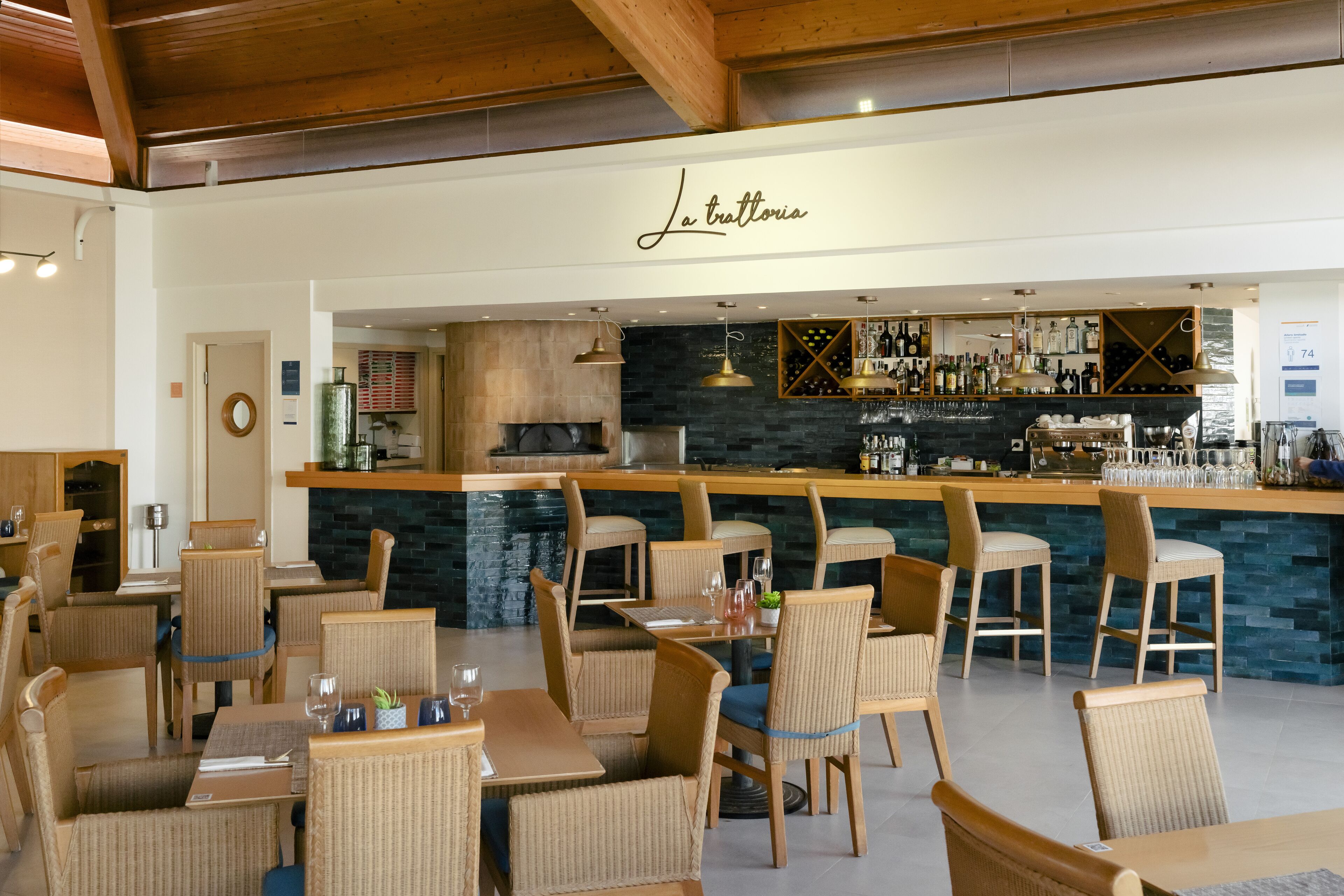 A restaurant with wicker chairs and tables, a bar area with wine bottles, and a sign reading "La trattoria".