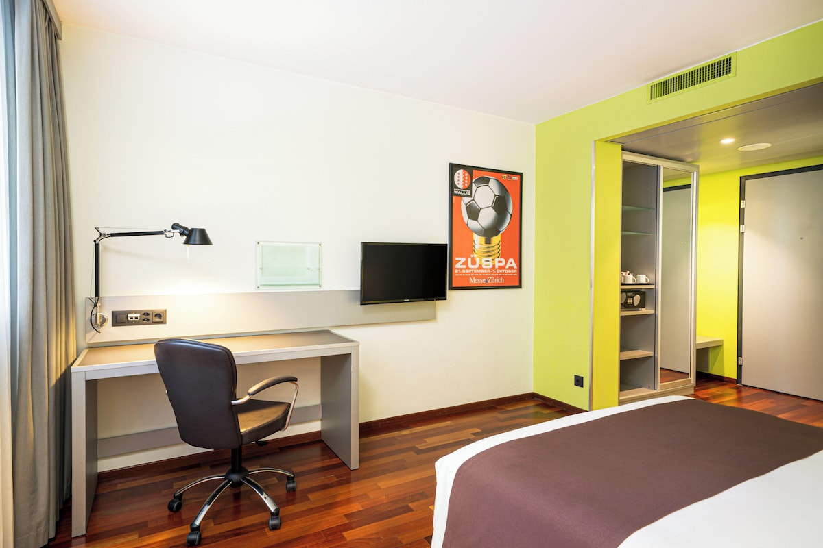 A hotel room with a bed, desk, chair, TV, and a poster on the wall.