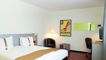 A hotel room with a bed, a desk, a chair, a mirror, and a poster on the wall.