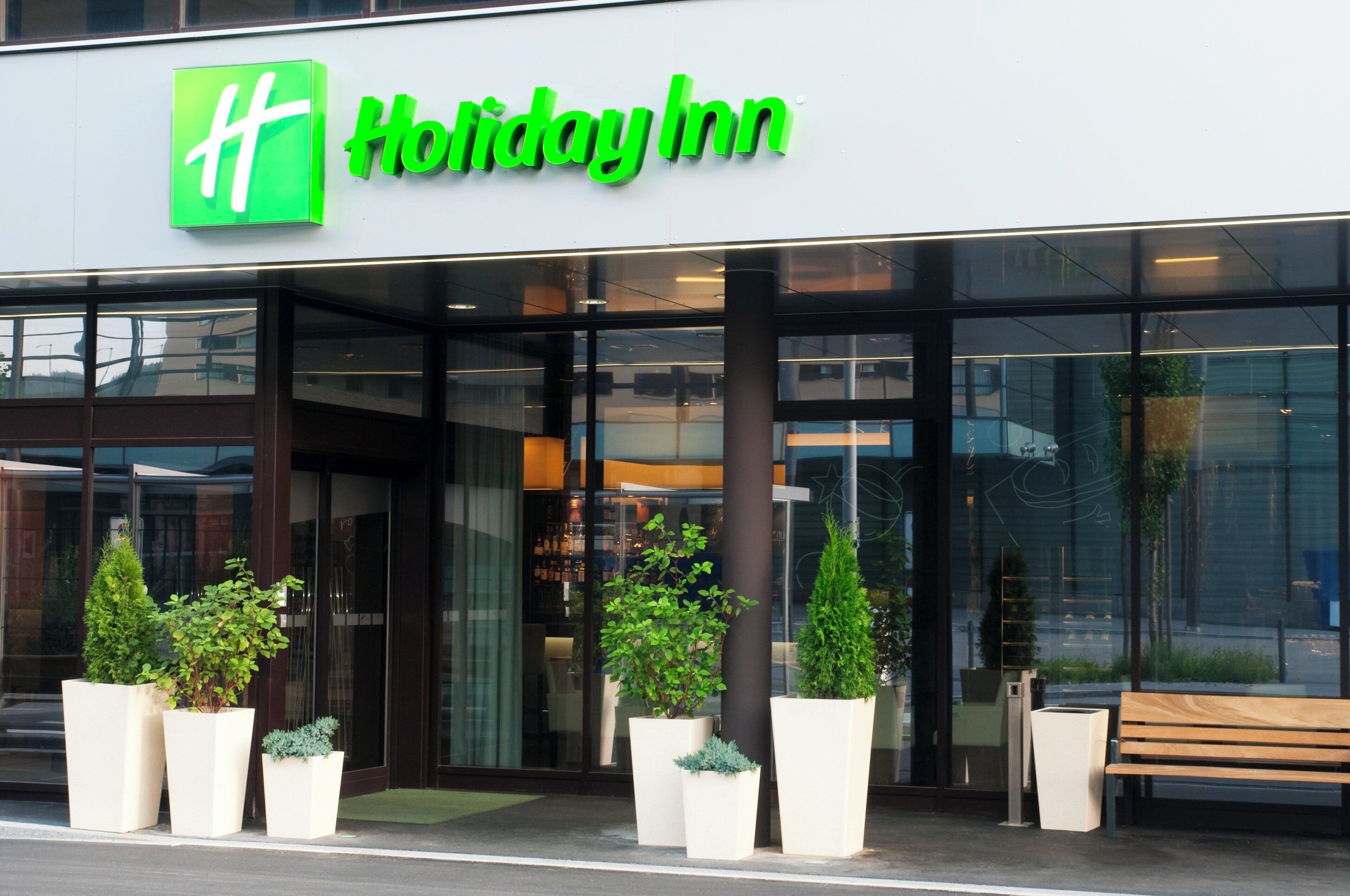 Holiday Inn -hotellin sisäänkäynti, jossa on suuret lasiovet, ruukuissa olevia kasveja ja ulkona penkki.
