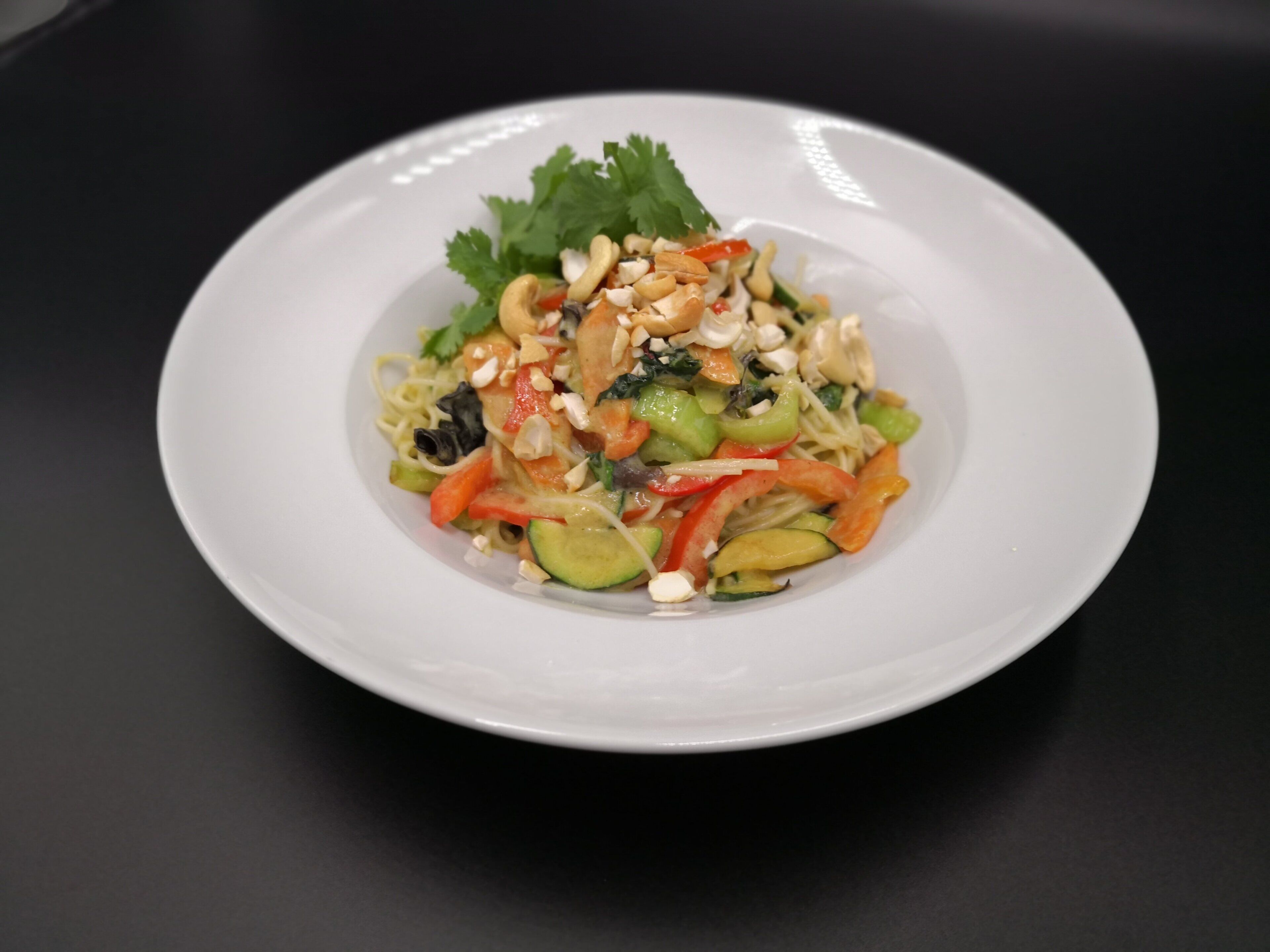 Un plato blanco con un plato de fideos, adornado con vegetales y nueces, sobre una superficie negra.