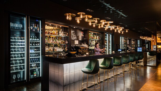 A bar area with a long counter, multiple stools, and shelves stocked with bottles and glasses. - Holiday Inn Zurich - Messe by IHG (Zürich)