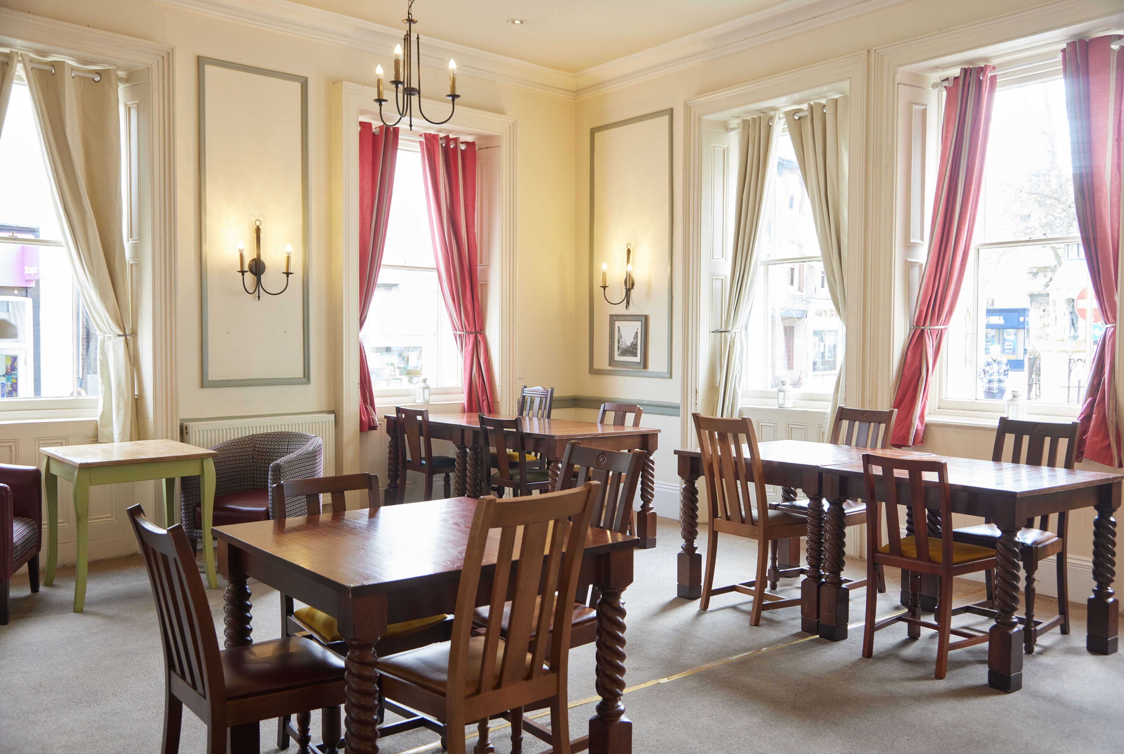 Un comedor con mesas y sillas de madera, ventanas amplias con cortinas, y lámparas fijadas a la pared.