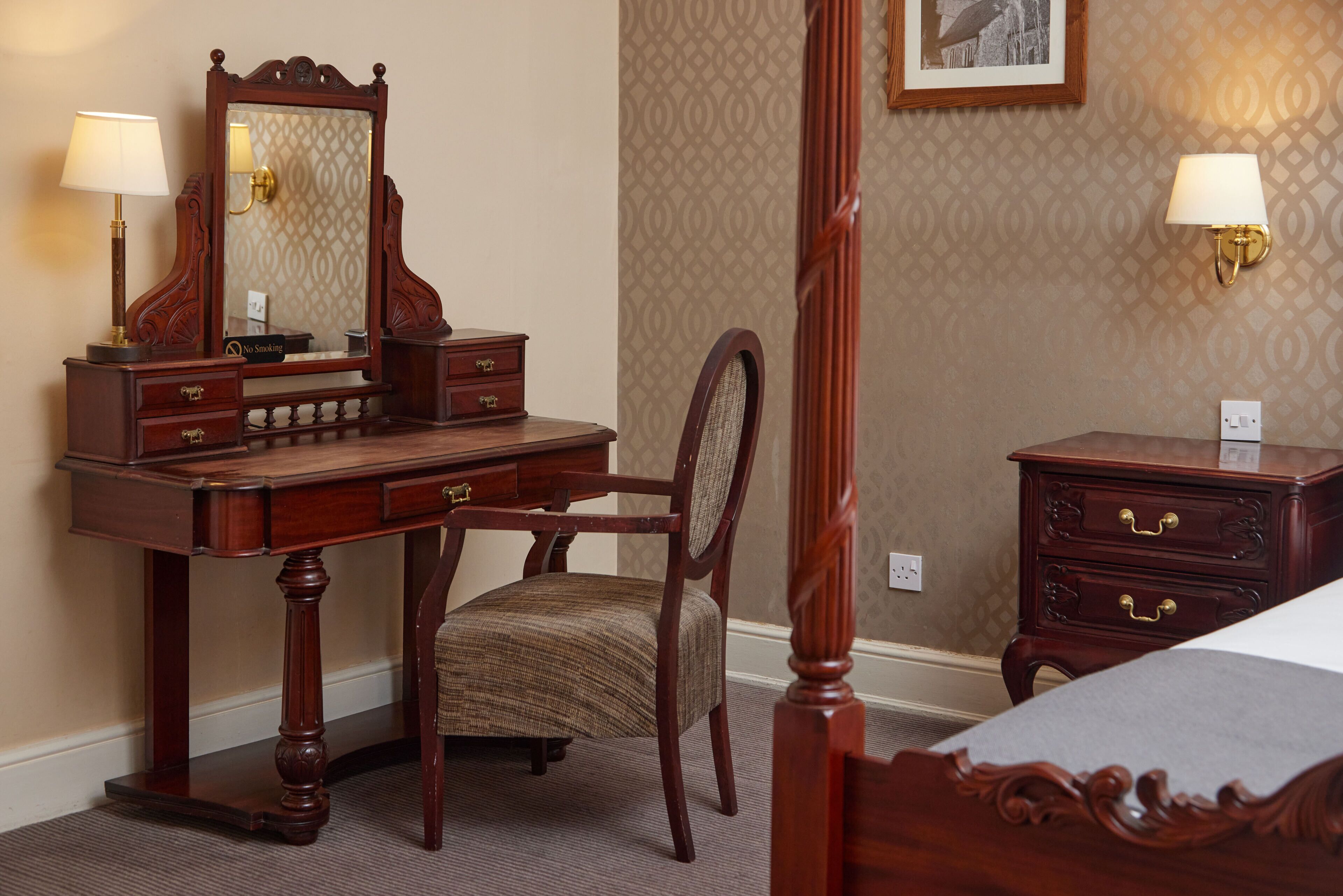 A bedroom with a wooden four-poster bed, a wooden desk with a chair, a dresser, and a nightstand with a lamp.