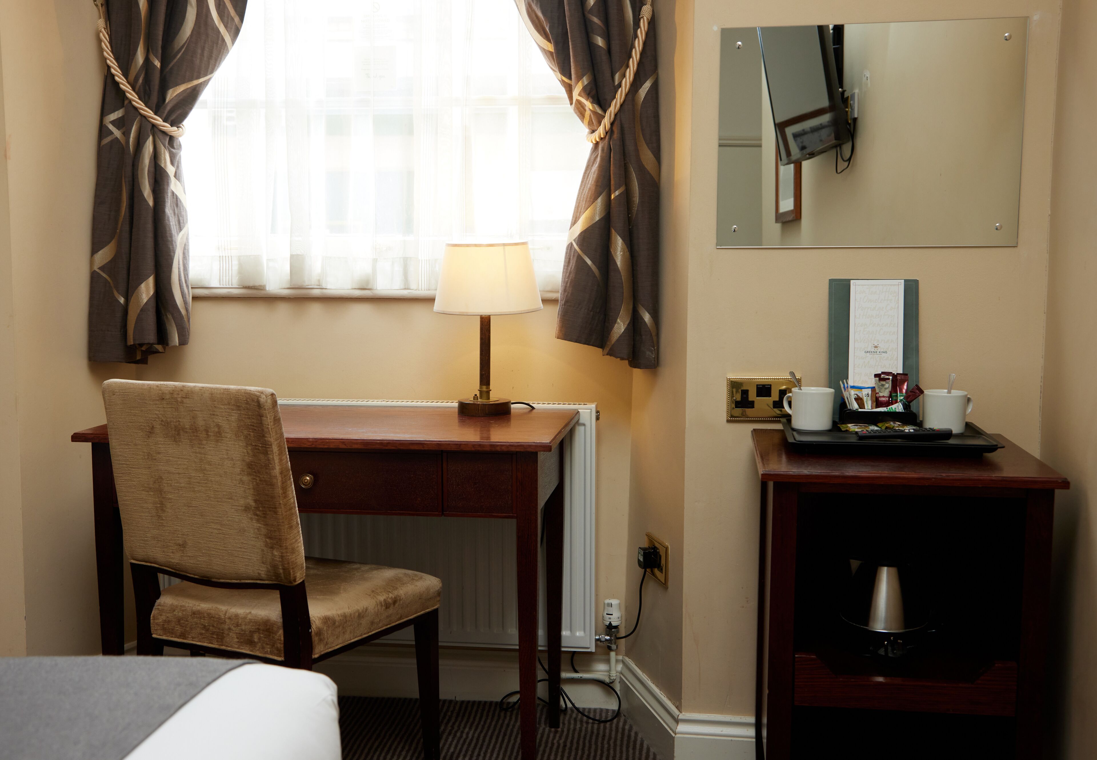 A hotel room with a bed, a desk with a lamp, a chair, a coffee machine, and a mirror.