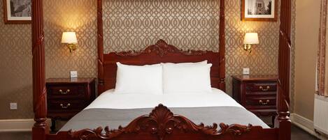 Ein Bett mit Baldachin, dessen Holzrahmen mit weißer Bettwäsche bezogen ist, mit Nachttischen mit Lampen an beiden Seiten, in einem Raum mit gemustertem Tapeten und gerahmten Bildern an der Wand.