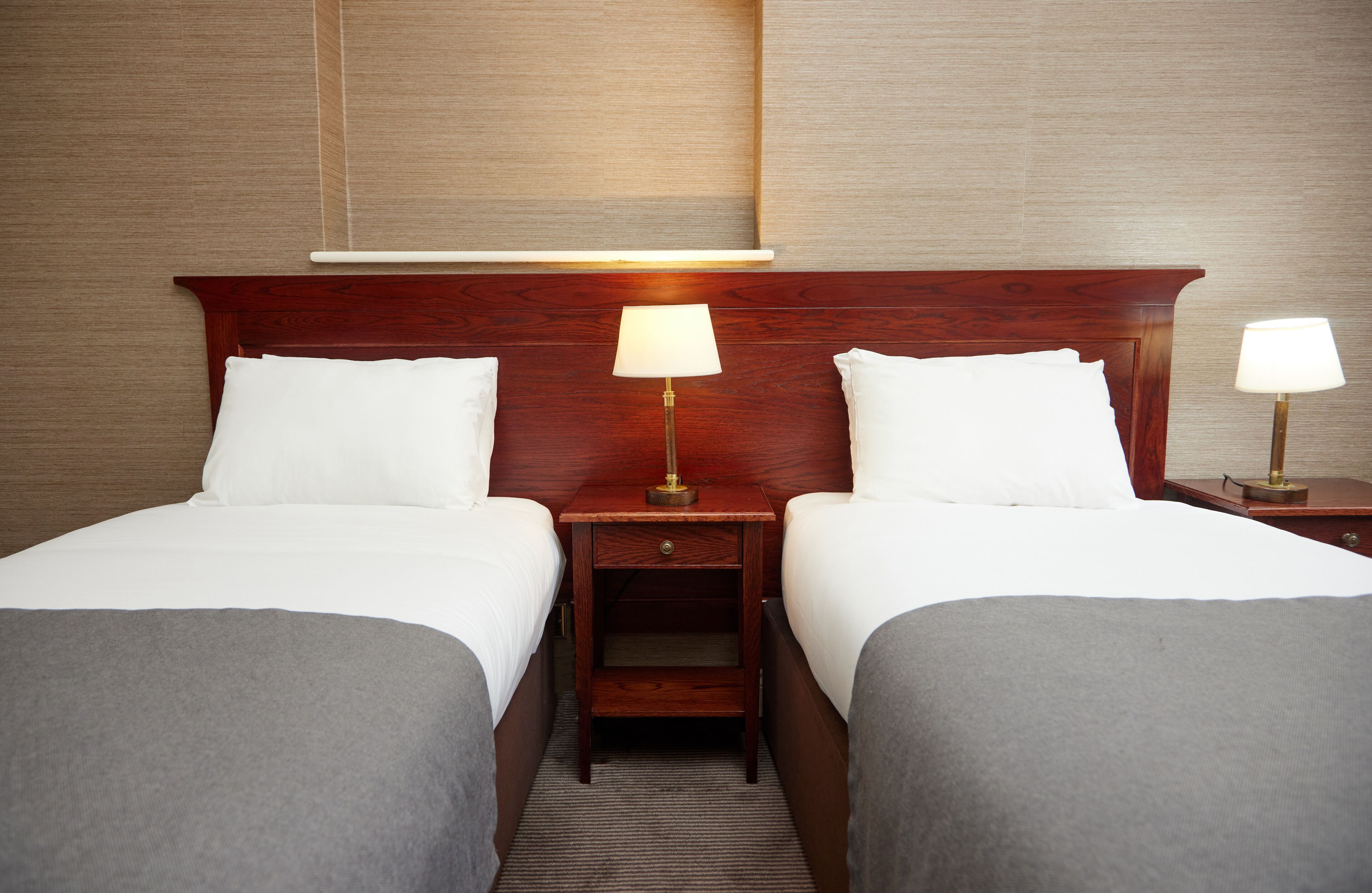 A hotel room with two single beds, wooden headboards, bedside tables with lamps, and a neutral color scheme.