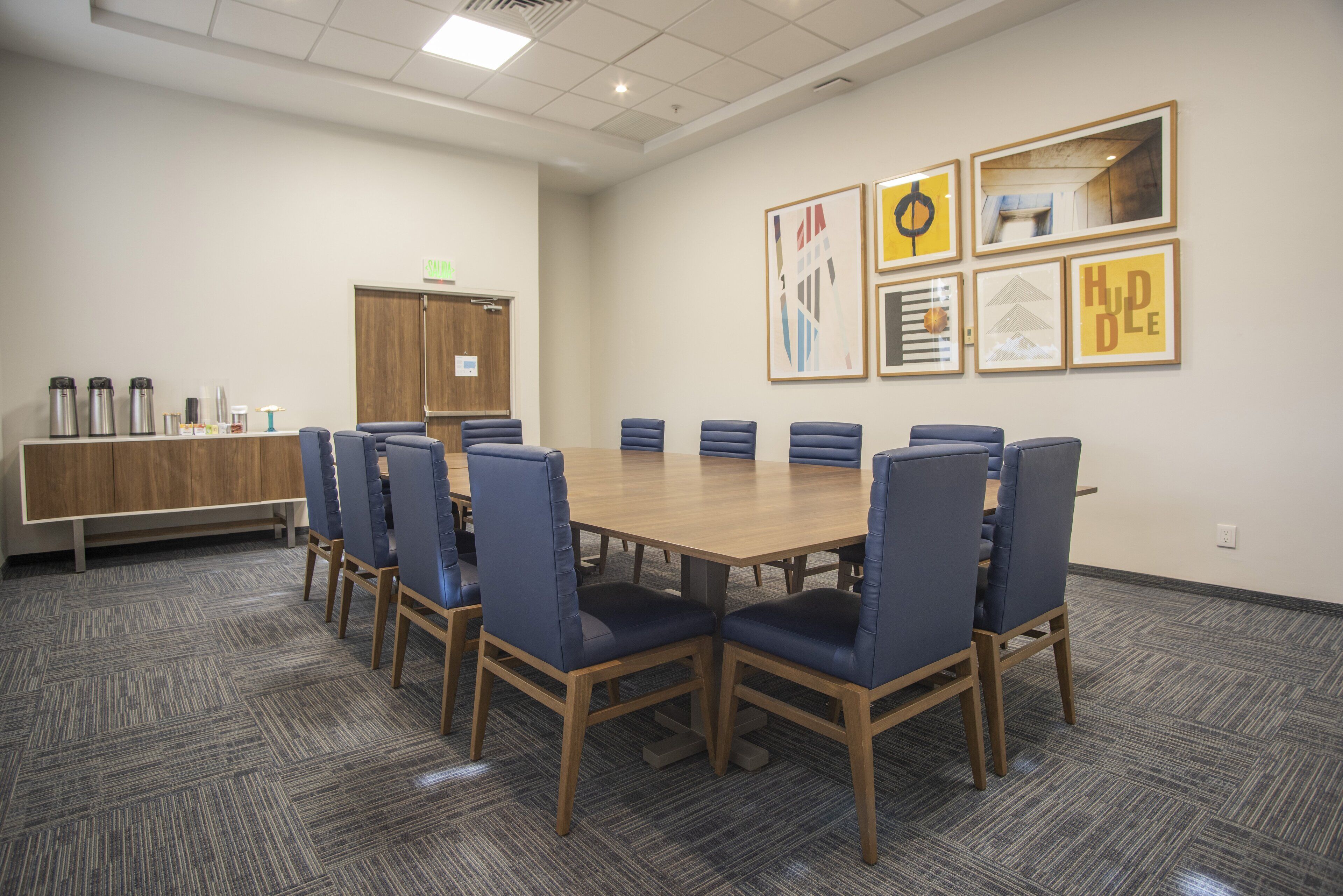Ein Konferenzraum mit einem langen Tisch, blauen Stühlen, einem Holzkasten und gerahmten Kunstwerken an der Wand.
