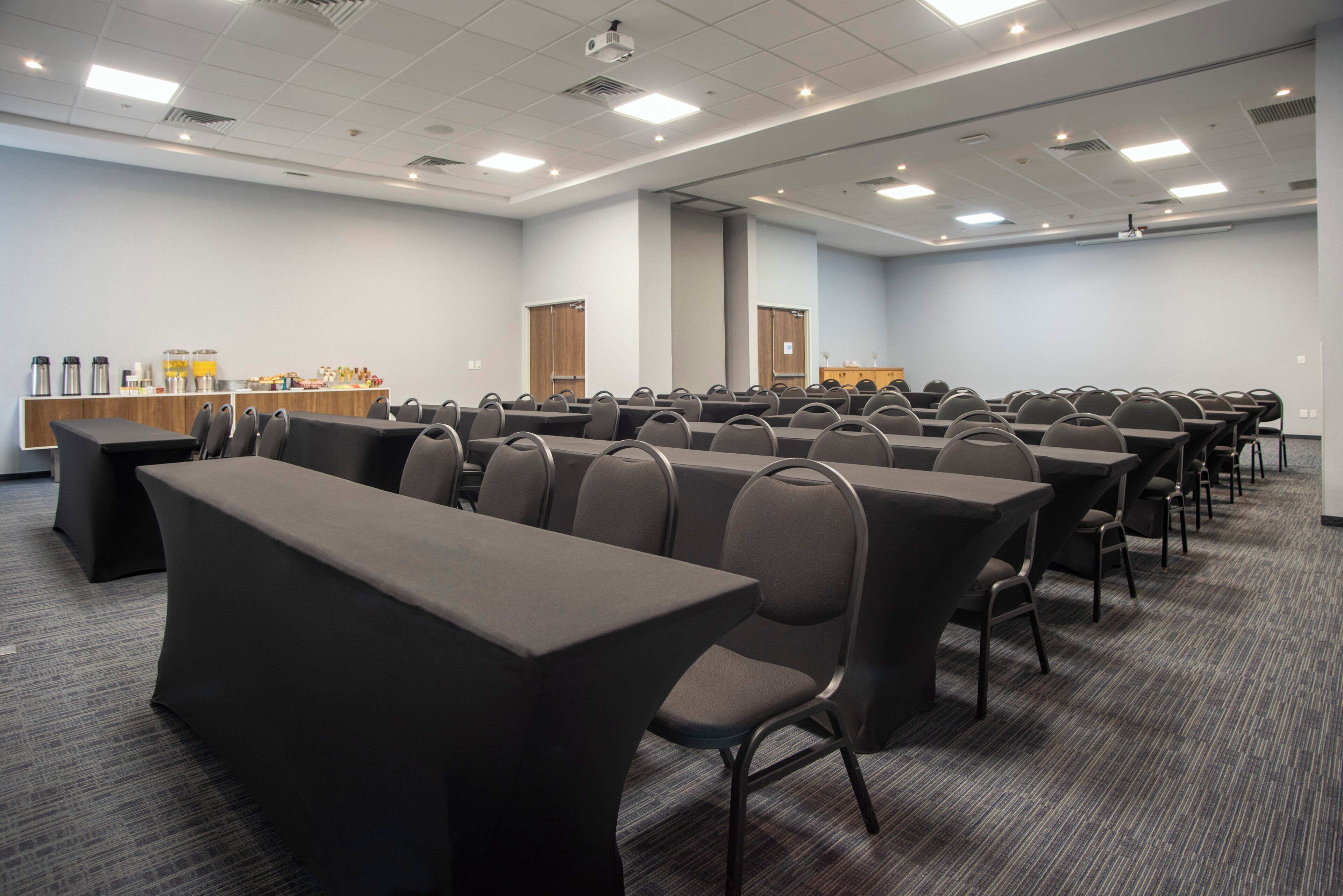 Een vergaderzaal met rijen tafels en stoelen, een buffetgedeelte en een projectiescherm.