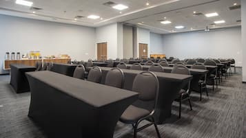 Een vergaderzaal met rijen tafels en stoelen, een buffetgedeelte en een projectiescherm.