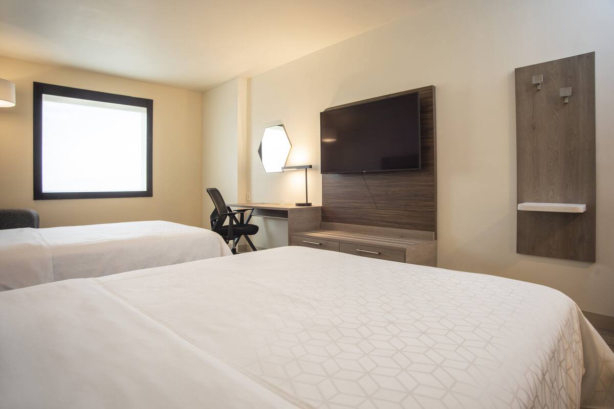 A hotel room with two beds, a desk, a chair, a TV, and a window.