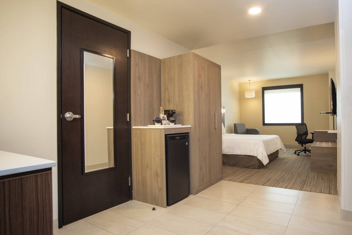 A hotel room with a wooden wardrobe, a small kitchenette, a bed, a desk, and a television.