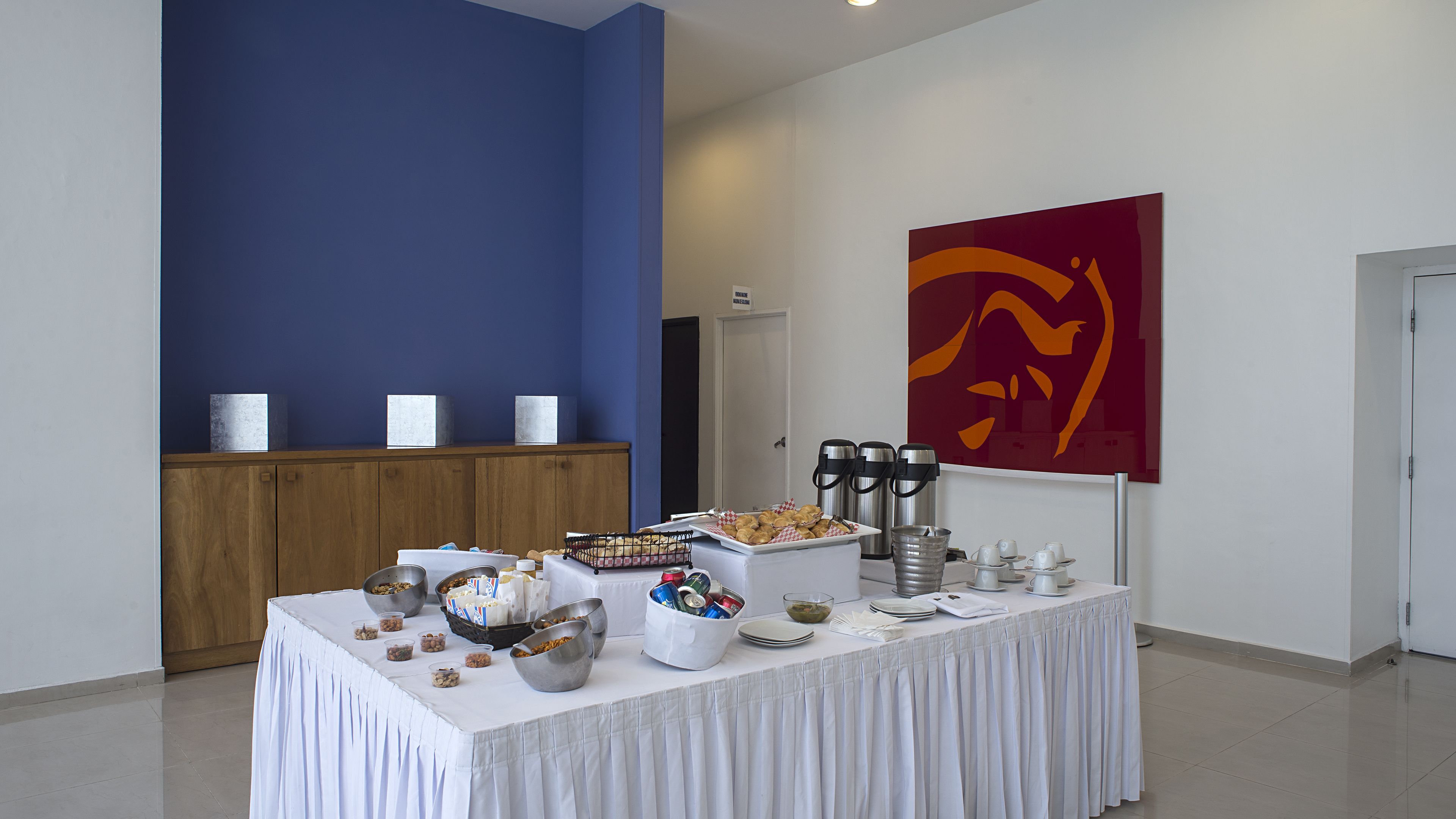 A buffet table with various food items and beverages, a wooden cabinet, and a large abstract painting on the wall.