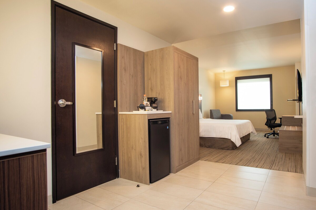 A hotel room with a wooden wardrobe, a small kitchenette, a bed, a desk, and a television.