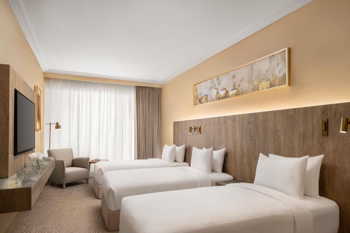Une chambre d’hôtel avec trois lits, un téléviseur à écran plat, un bureau avec une lampe et une grande fenêtre avec des rideaux.