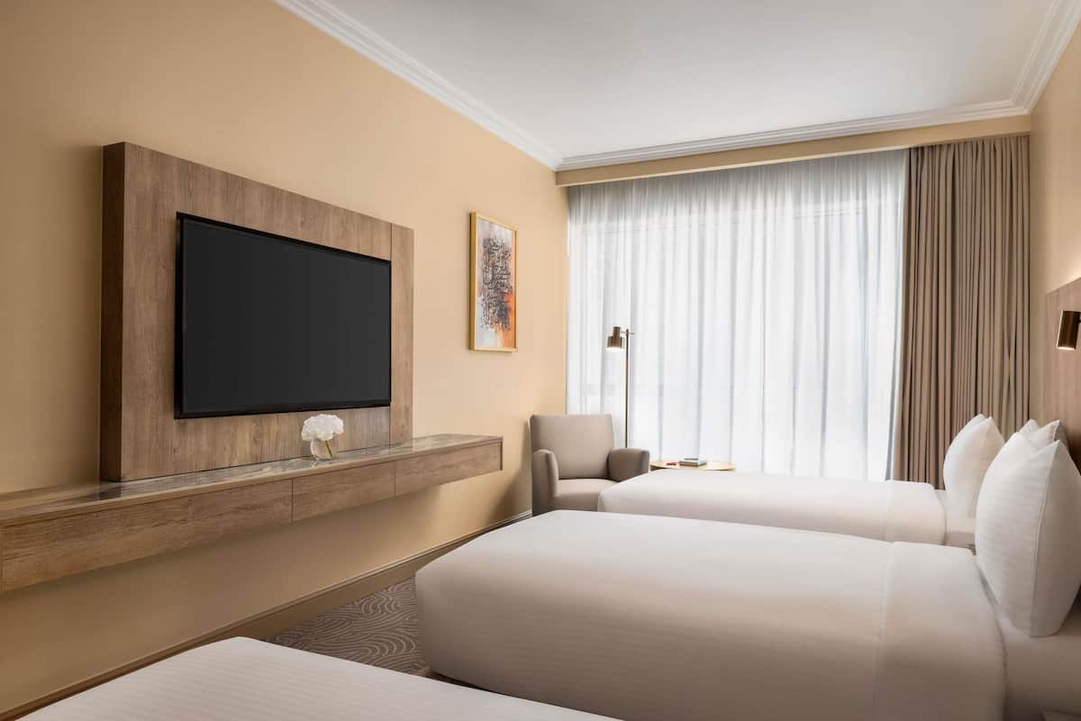 Een hotelkamer met twee bedden, een televisie bevestigd aan een houten paneel, een bijzettafeltje met een vaas en een groot raam met gordijnen.