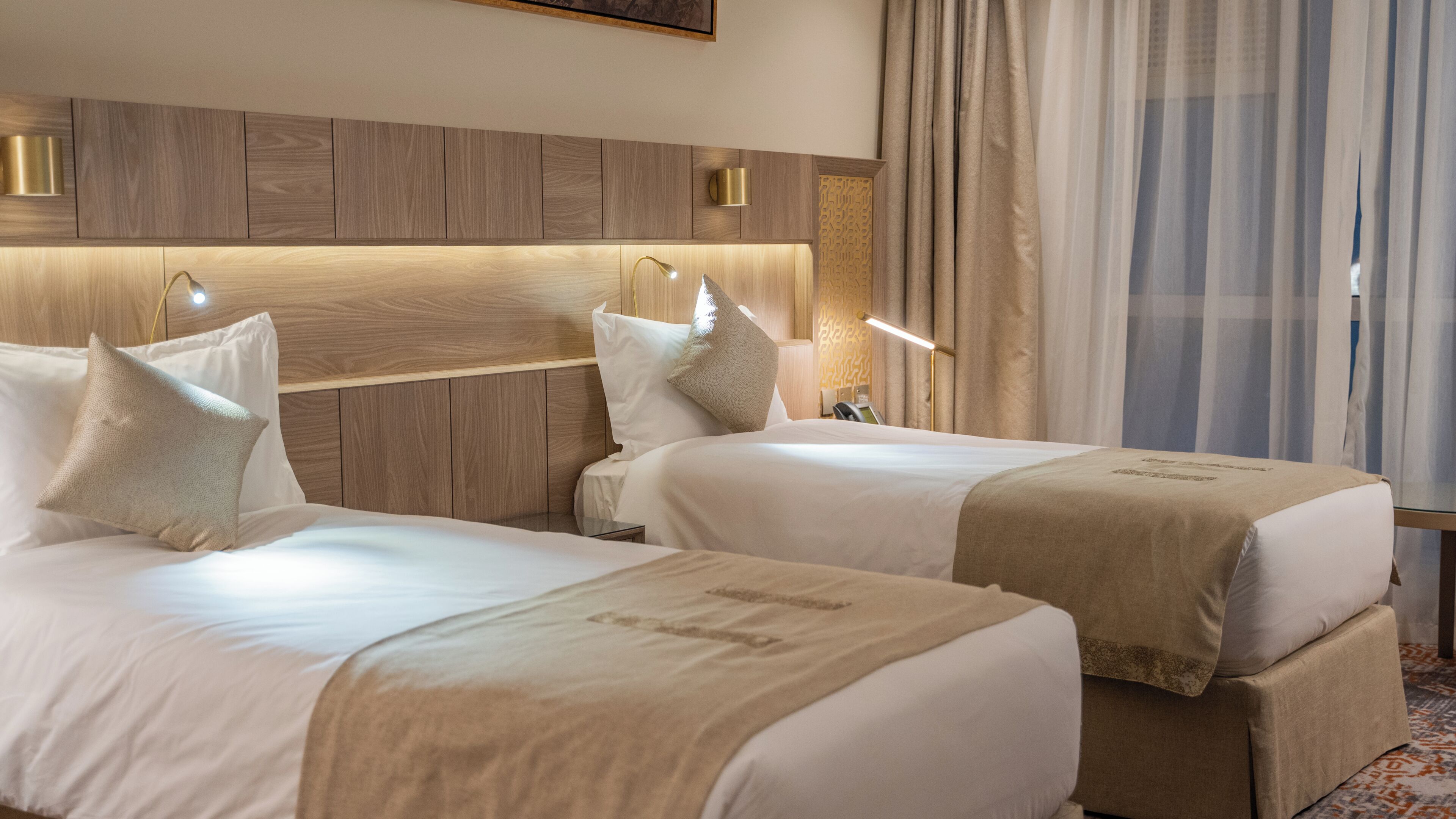 Ein Hotelzimmer mit zwei Betten, einem Holzkopfende, Nachttischlampen und einem Fenster mit Vorhängen.
