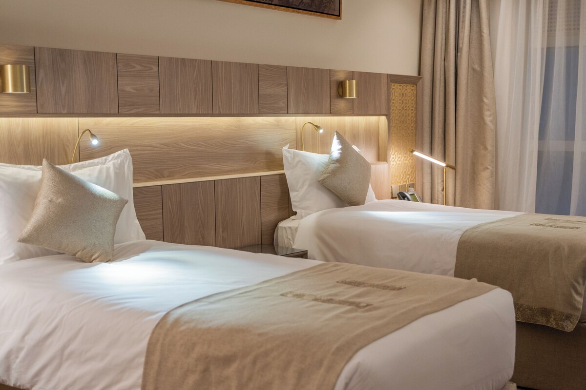 A hotel room with two beds, a wooden headboard, bedside lamps, and a window with curtains.