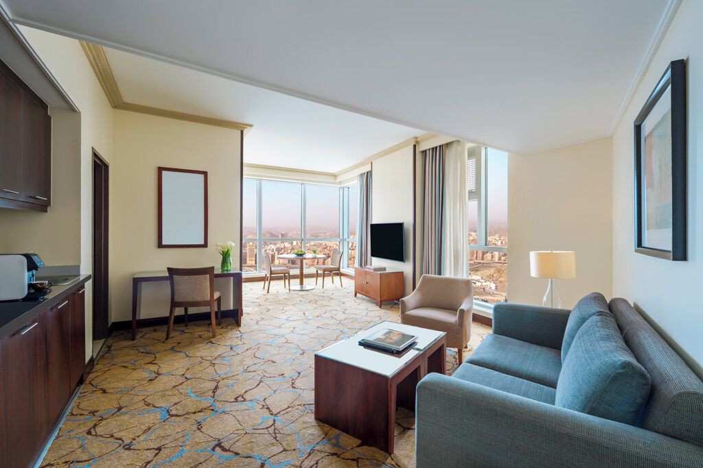 A hotel room with a large window, a sofa, a coffee table, a TV, and a dining area with a table and chairs.