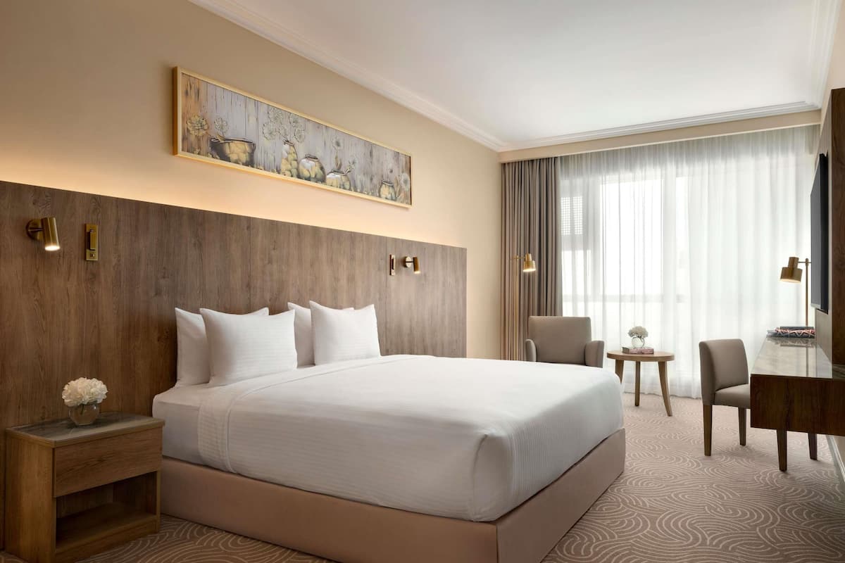 Une chambre d’hôtel avec un grand lit, une tête de lit en bois, un tableau au-dessus du lit, un bureau avec une chaise et une grande fenêtre avec des rideaux.