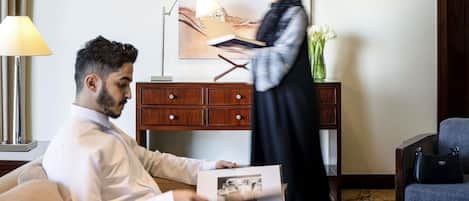 A hotel room with a sofa, a wooden side table, a lamp, a painting, and a vase with flowers.