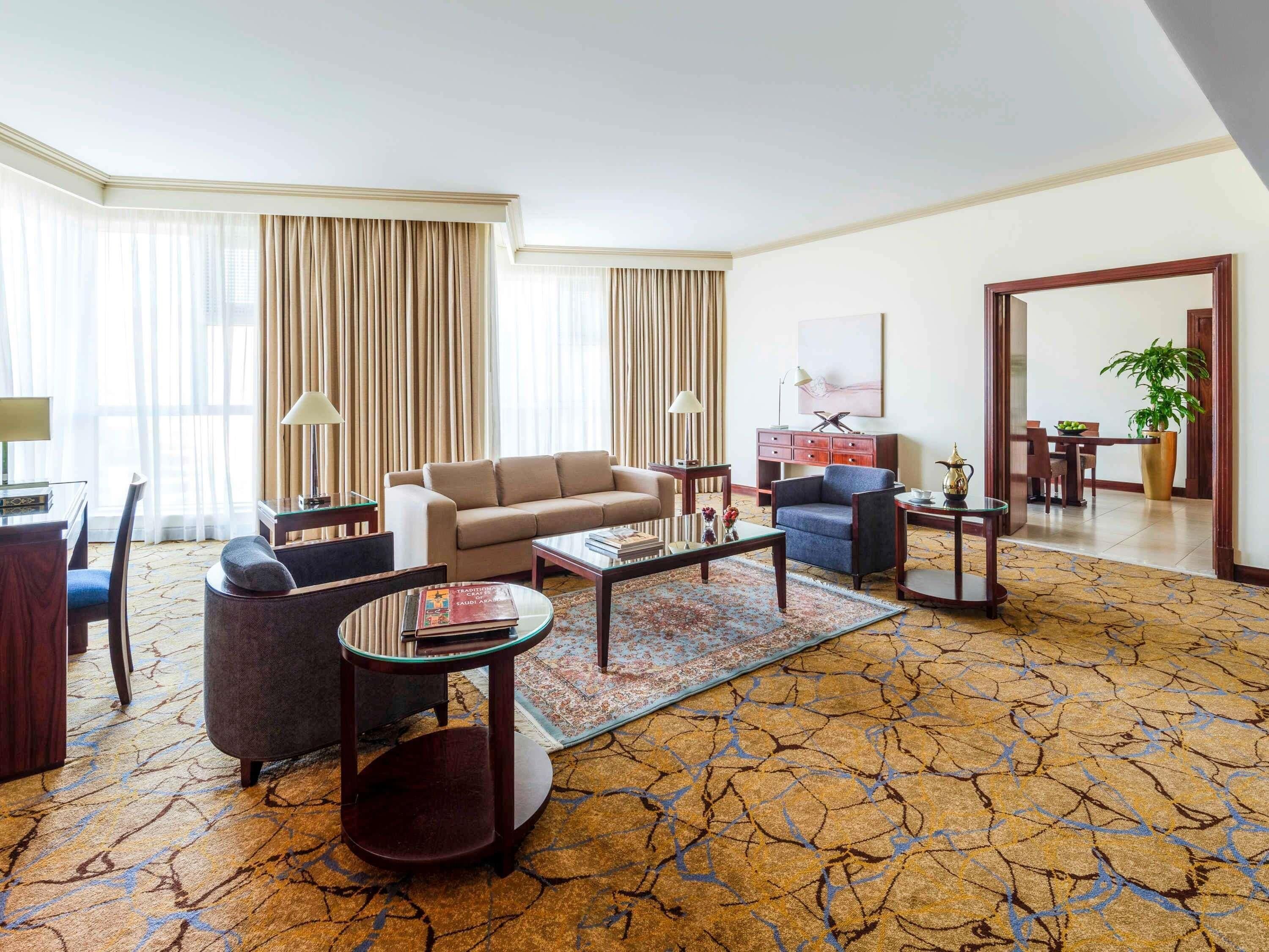 A hotel room with a sofa, armchairs, a coffee table, a desk, and a dining area.