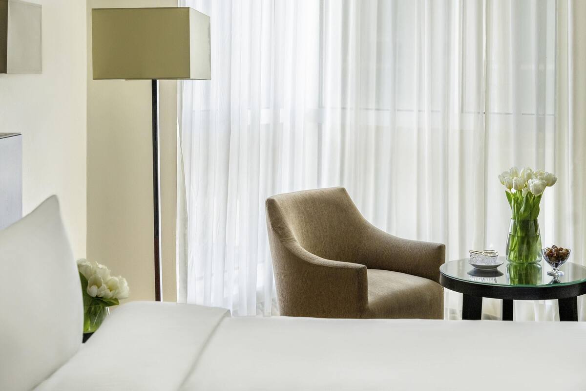 Een hotelkamer met een bed, een stoel, een tafel met een bloevas en een lamp.