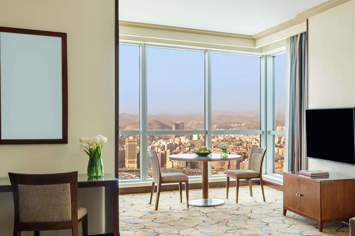 A room with a large window, a round table, two chairs, a cabinet, and a flat-screen TV.
