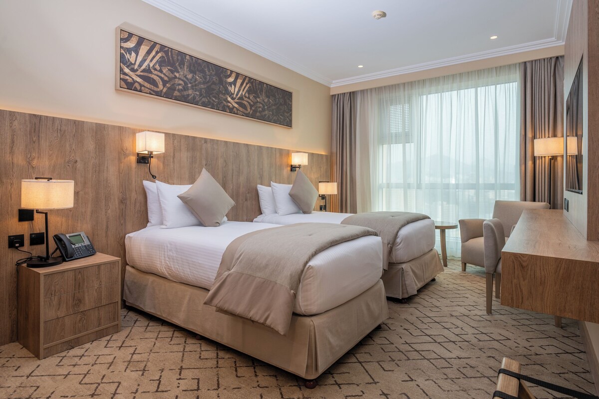 Une chambre d’hôtel avec deux lits, un bureau, une chaise, un téléphone et une grande fenêtre avec des rideaux.