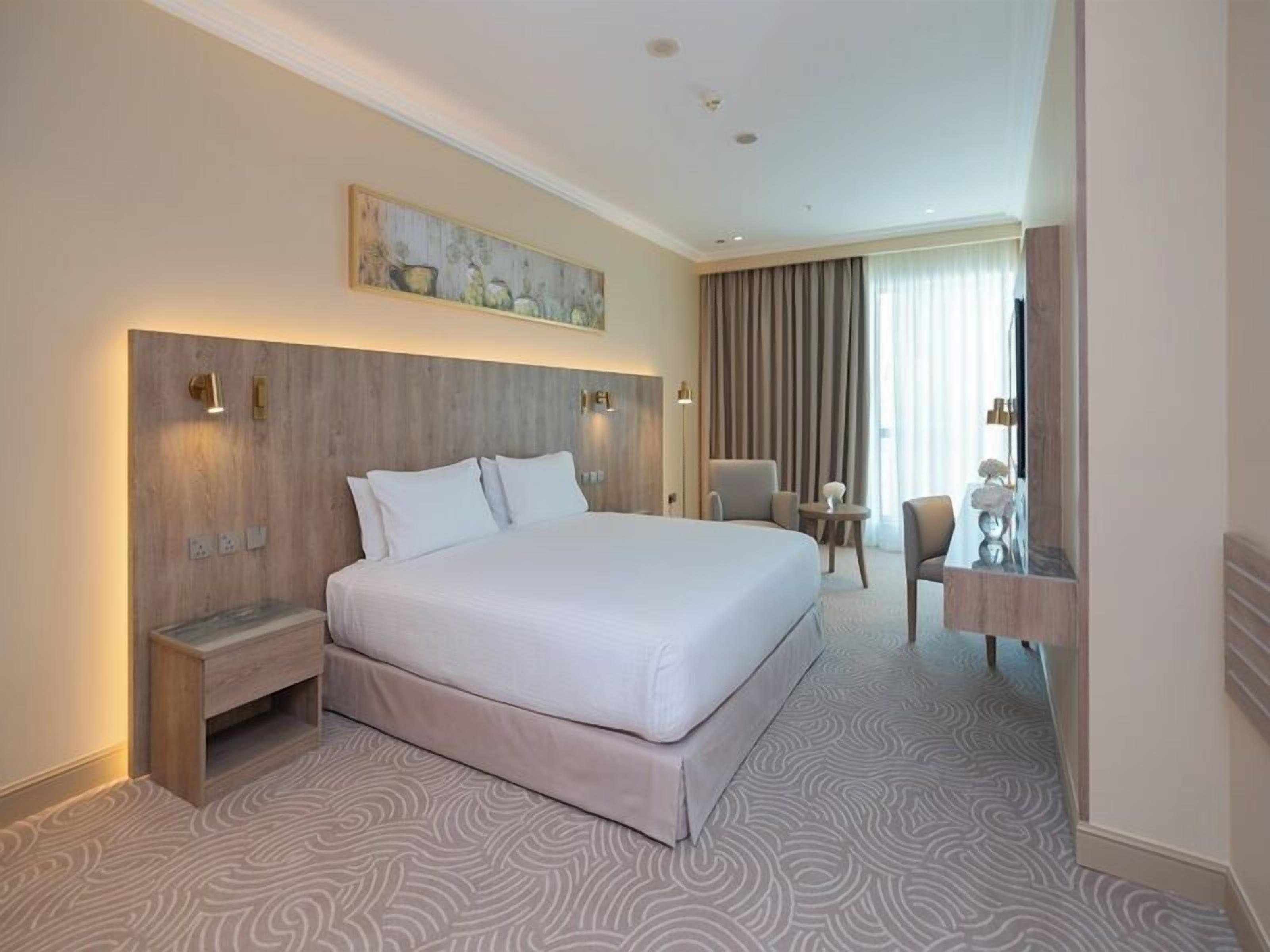 A hotel room with a large bed, bedside tables, a desk, a chair, a mirror, and a painting on the wall.