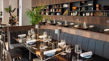 A modern restaurant with a long dining table, cushioned seating, and a variety of decorative items on shelves.