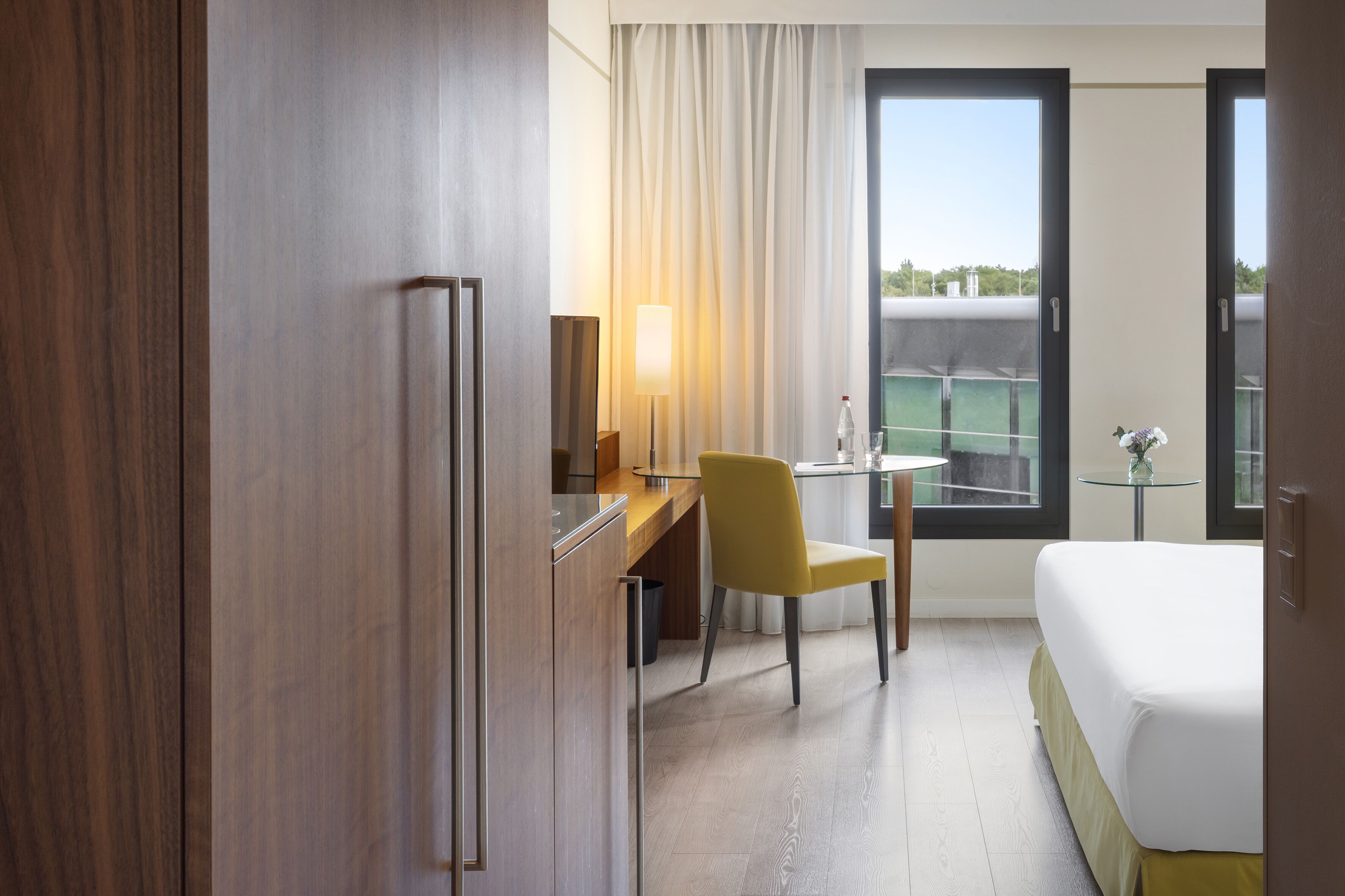 A hotel room with a bed, a desk, a chair, a lamp, a small table, and a view of the outdoors.
