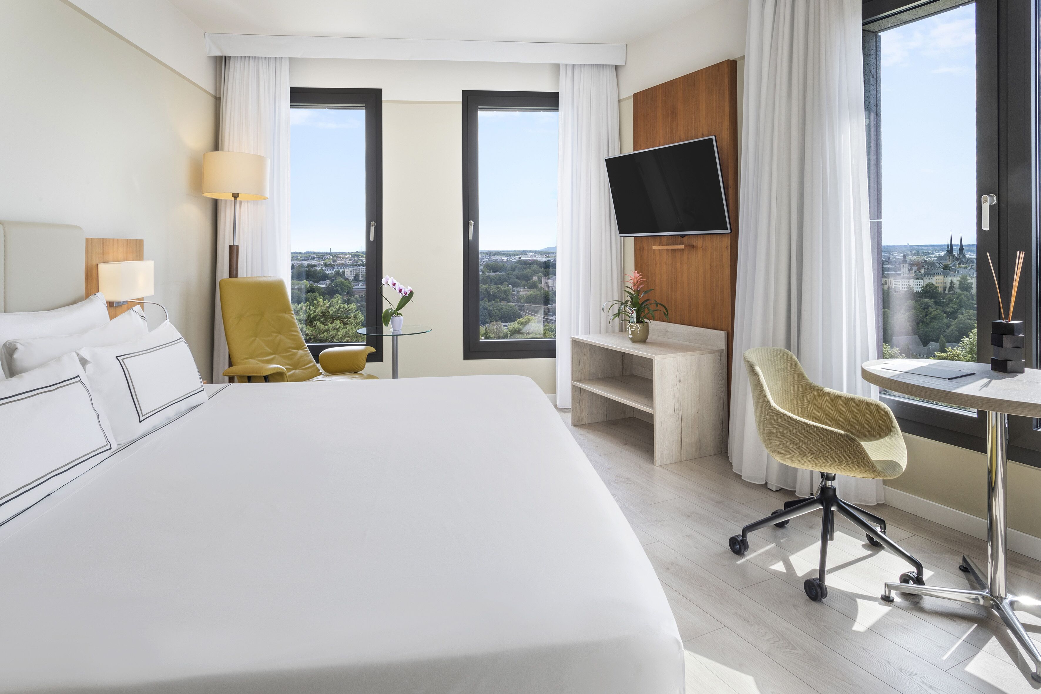 A hotel room with a large bed, a desk with a chair, a TV, and a view of the city through large windows.