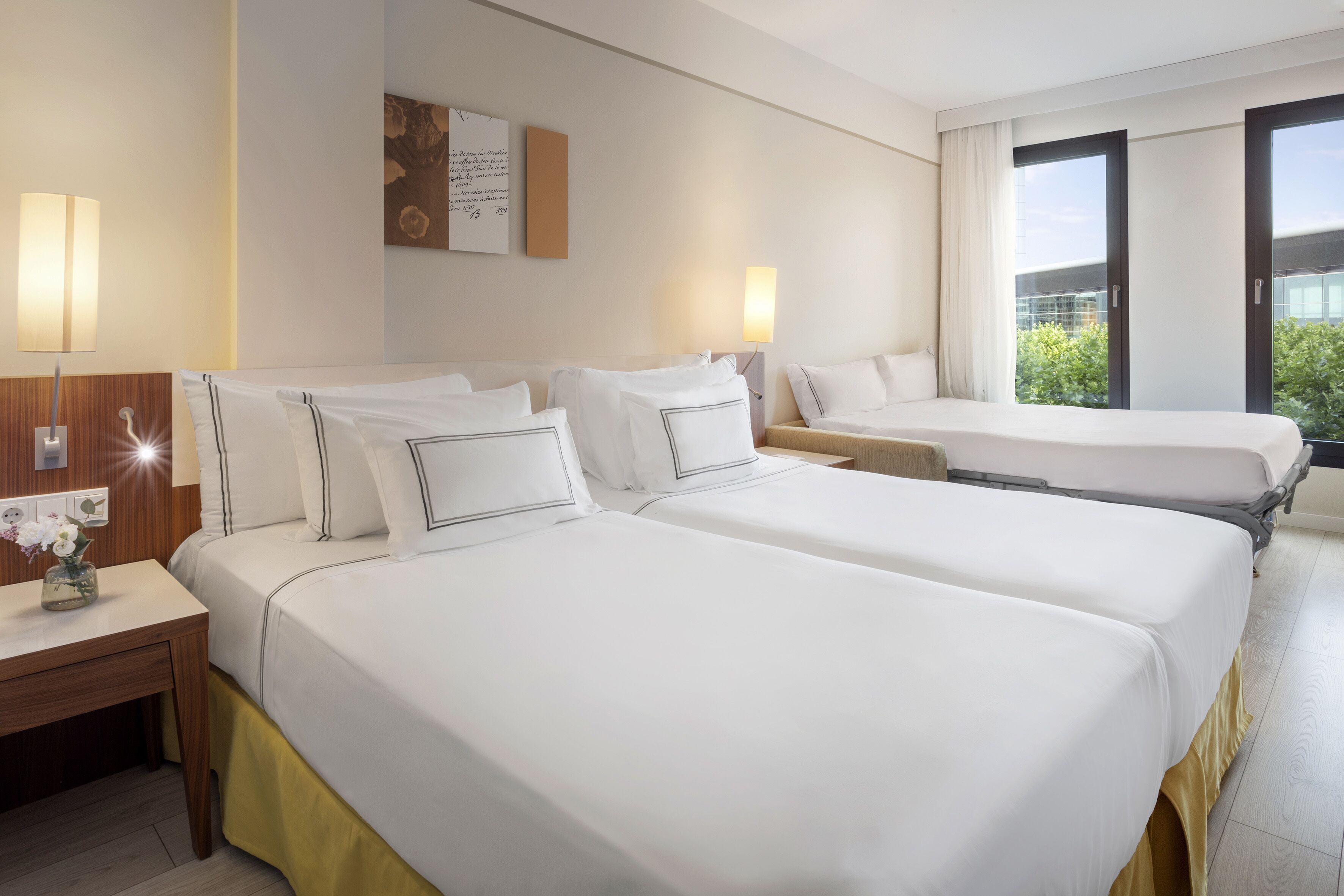 A hotel room with two beds, a wooden nightstand, a small table with a vase of flowers, and a large window with a view of greenery.