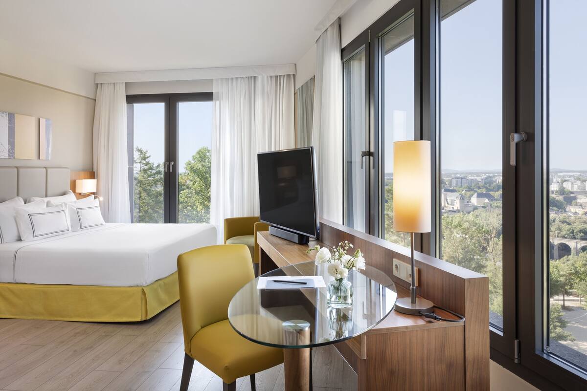 Una habitación de hotel con una cama, un escritorio con televisor, una mesa de vidrio y vistas a la ciudad.
