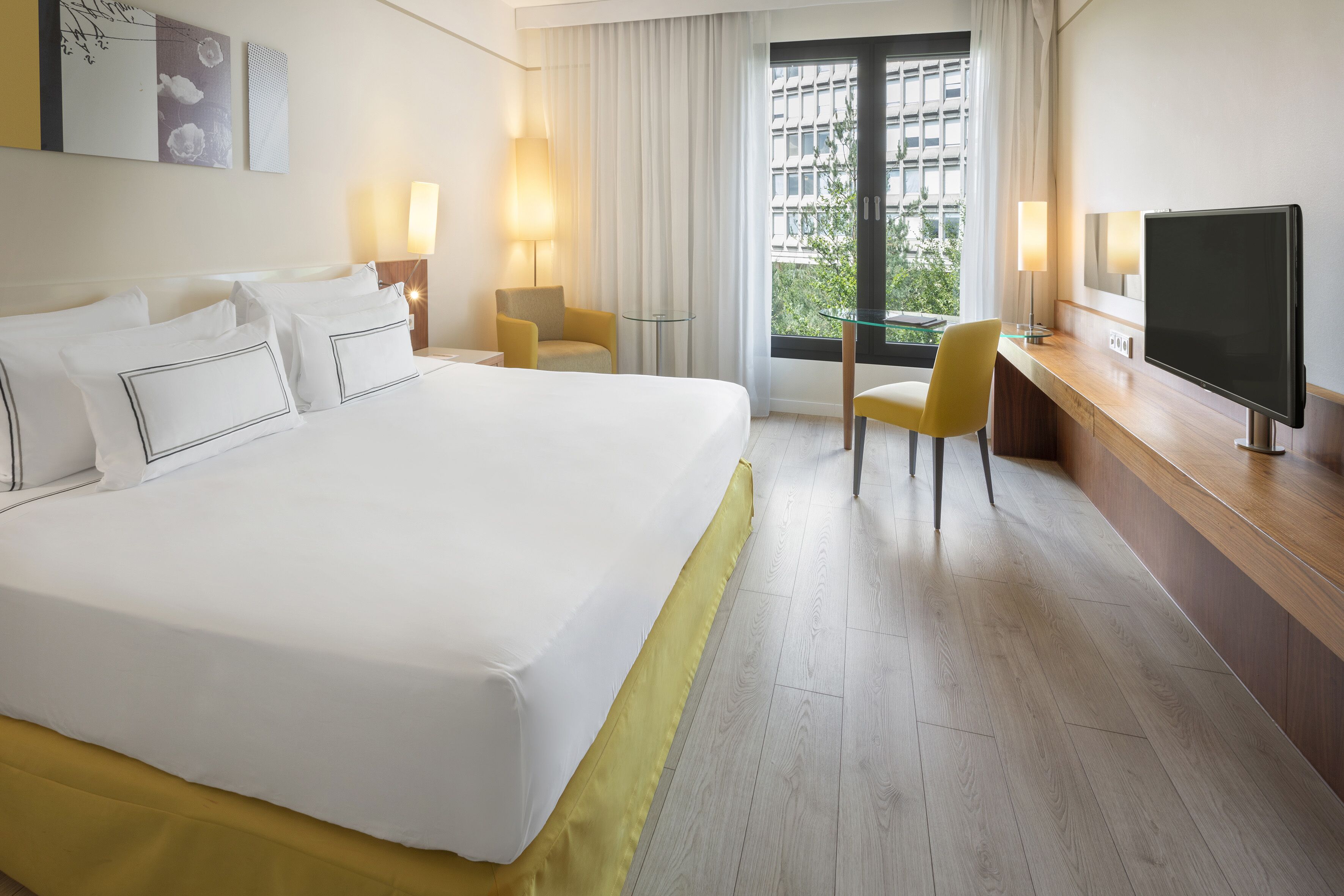 Una habitación de hotel con una cama grande, un escritorio con televisor, dos sillas y un ventanal con cortinas.