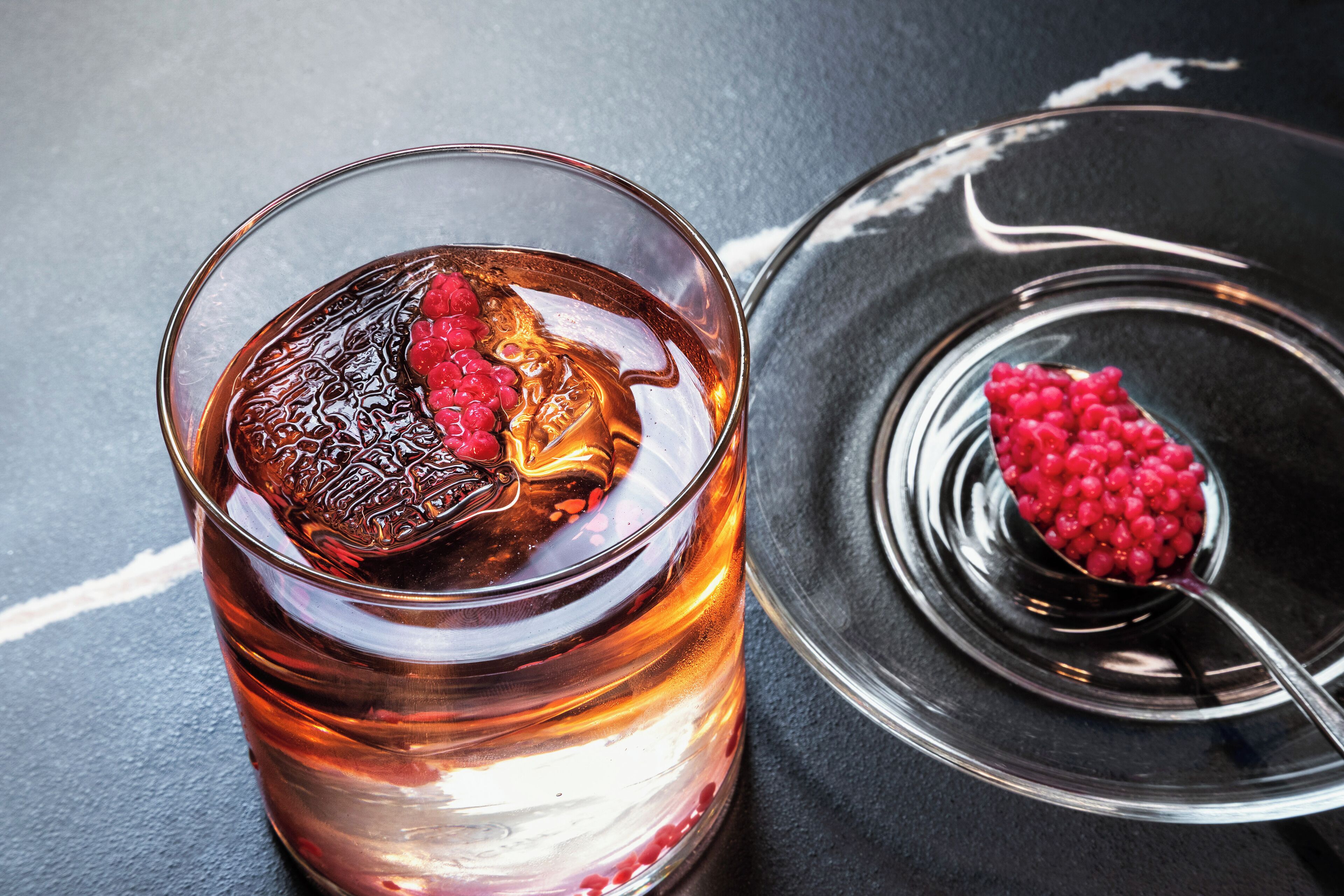 Een glas amberkleurig drankje met ijs en een plakje rood fruit, met een lepel rode bessen op een reflecterend oppervlak.
