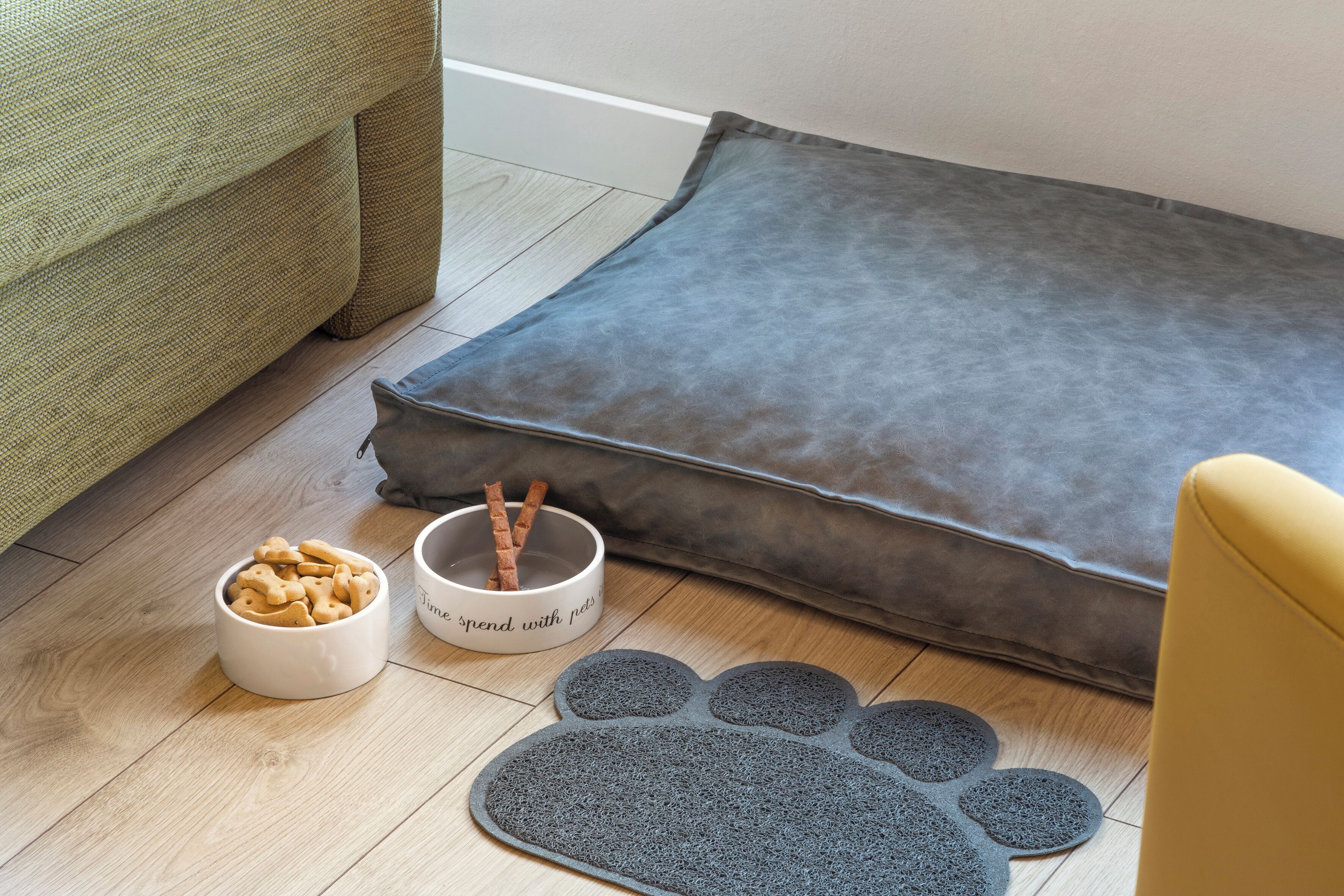 A pet bed, a bowl of pet treats, and a pet paw print mat on a wooden floor.
