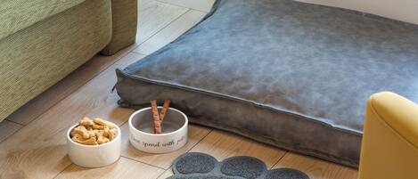 A pet bed, a bowl of pet treats, and a pet paw print mat on a wooden floor.