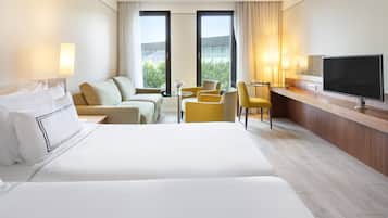 Habitación de hotel con una cama grande, un sofá, un escritorio con televisor y dos ventanas con cortinas.