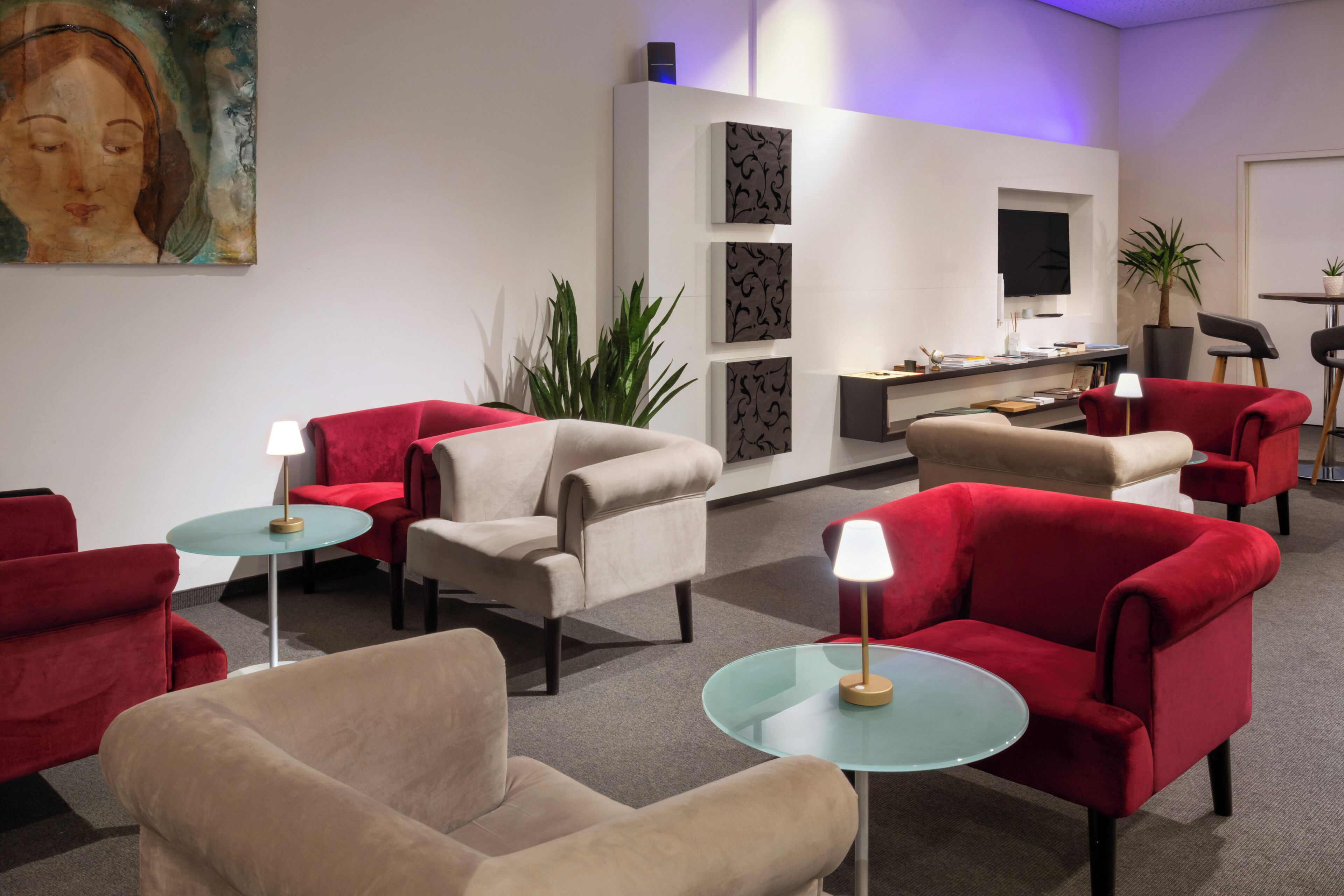 Un moderno vestíbulo de hotel con sillones rojos y beiges, una mesa de centro de vidrio, un escritorio con una computadora y una planta en maceta.