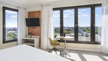 A modern hotel room with a large bed, a TV mounted on the wall, a small desk with a chair, and a large window offering a city view.