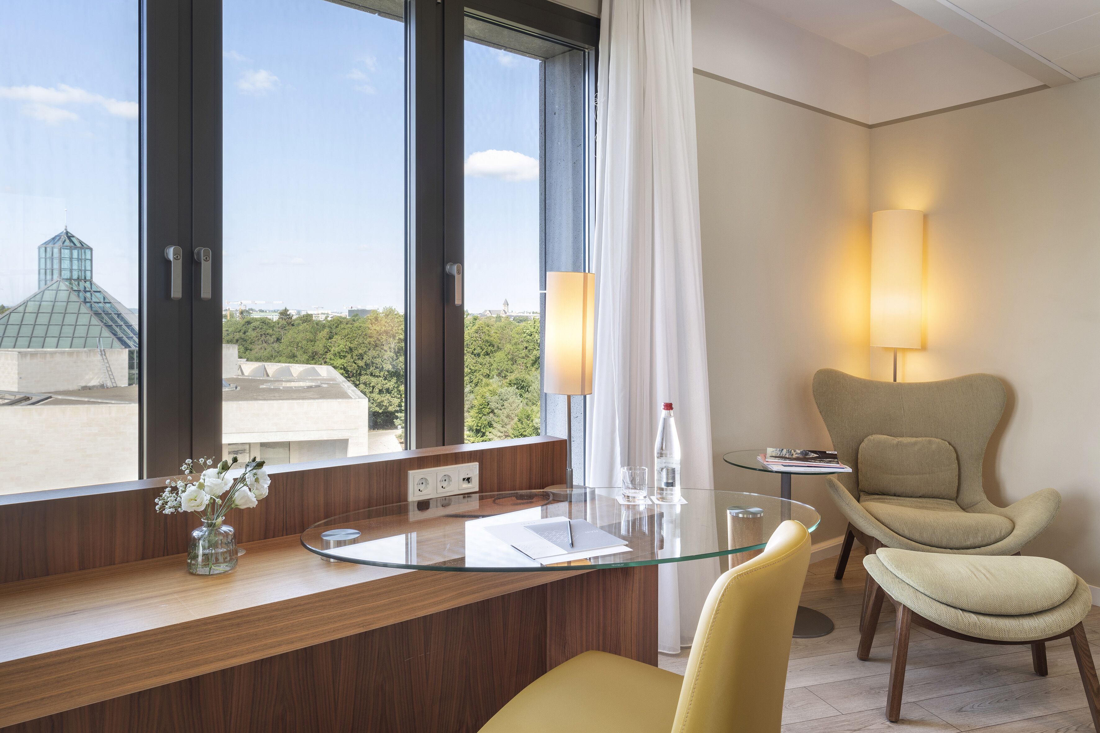 Una habitación de hotel moderna con un escritorio de cristal, una silla amarilla, un sillón beige, una lámpara y vistas a la ciudad.
