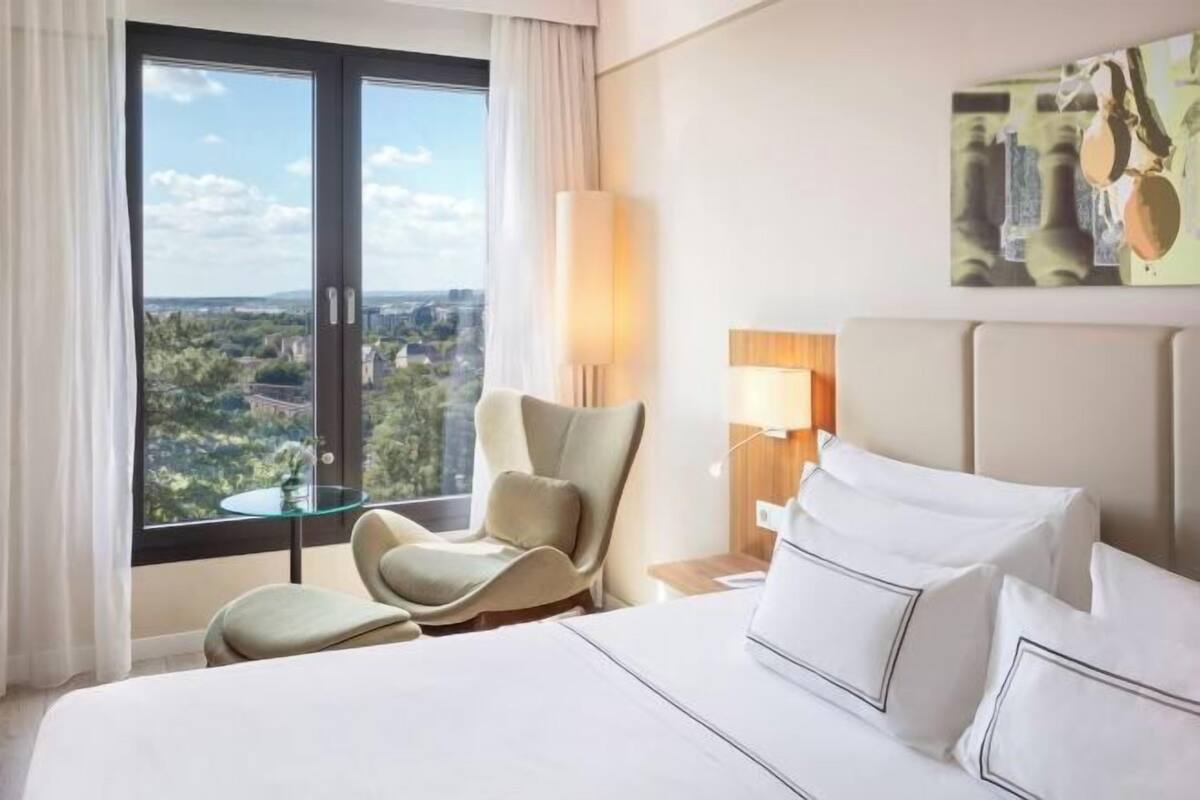 Habitación de hotel con cama, una silla, una mesa y un amplio ventanal con vistas a la ciudad.