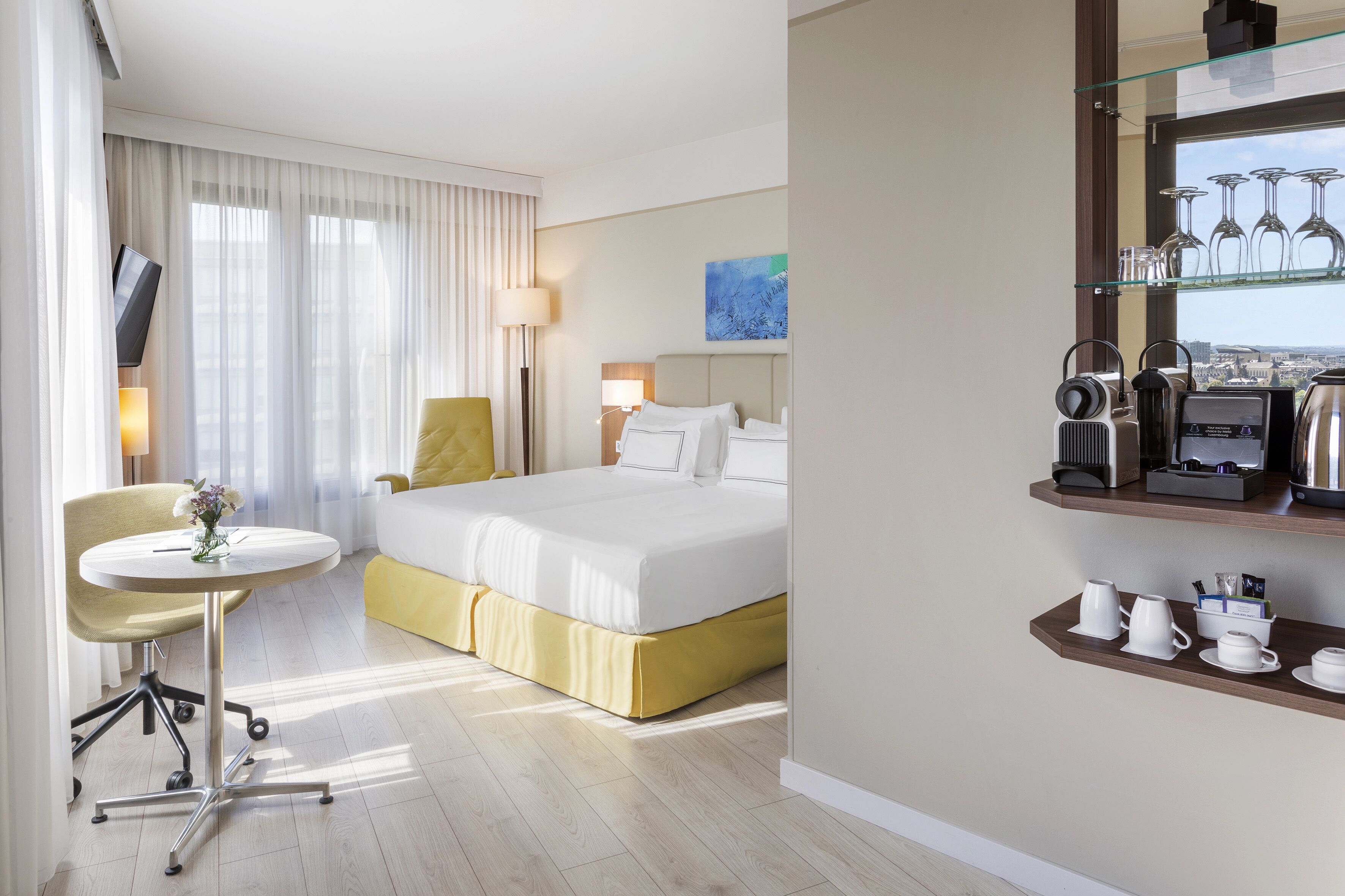 Une chambre d’hôtel comprenant un lit, un bureau, une chaise, une petite table avec un vase, une machine à café et une étagère avec des tasses et des verres.