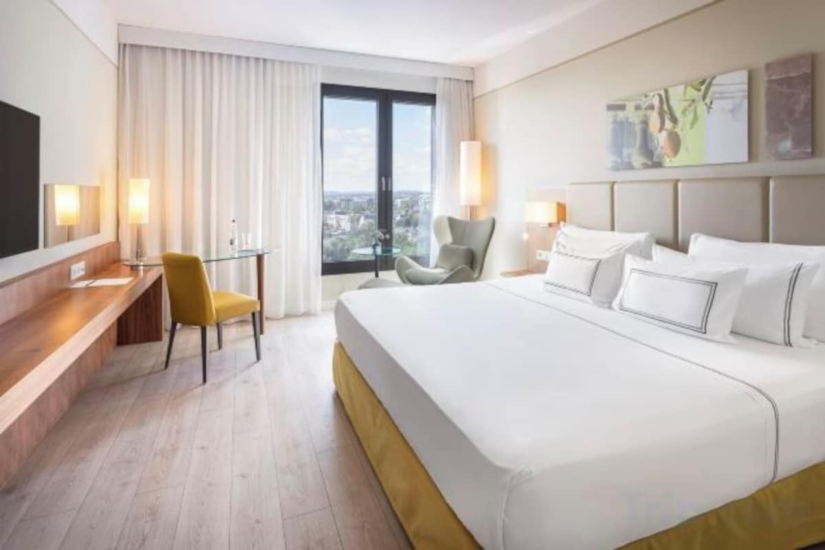 A hotel room with a large bed, a desk, two chairs, a TV, and a window with curtains.