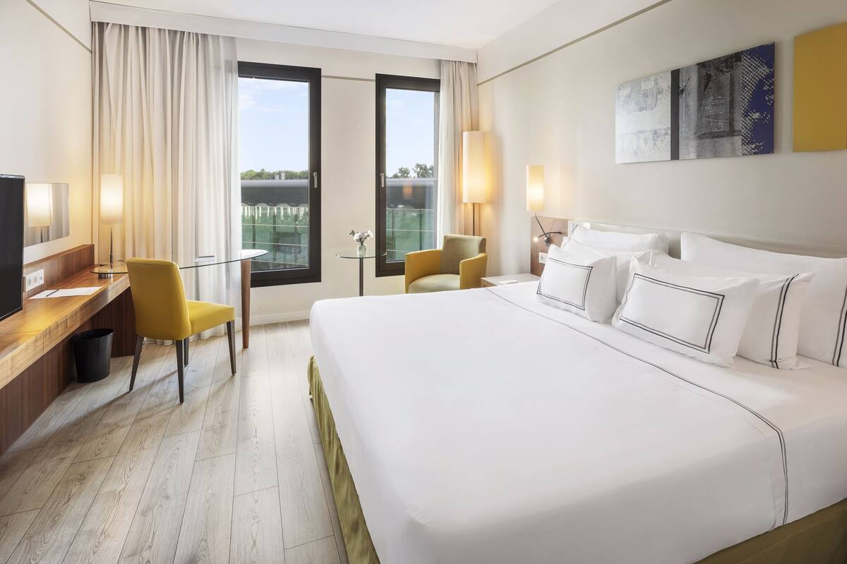 Una habitación de hotel con una cama grande, un escritorio con una silla, una mesita de noche con una lámpara y un televisor.