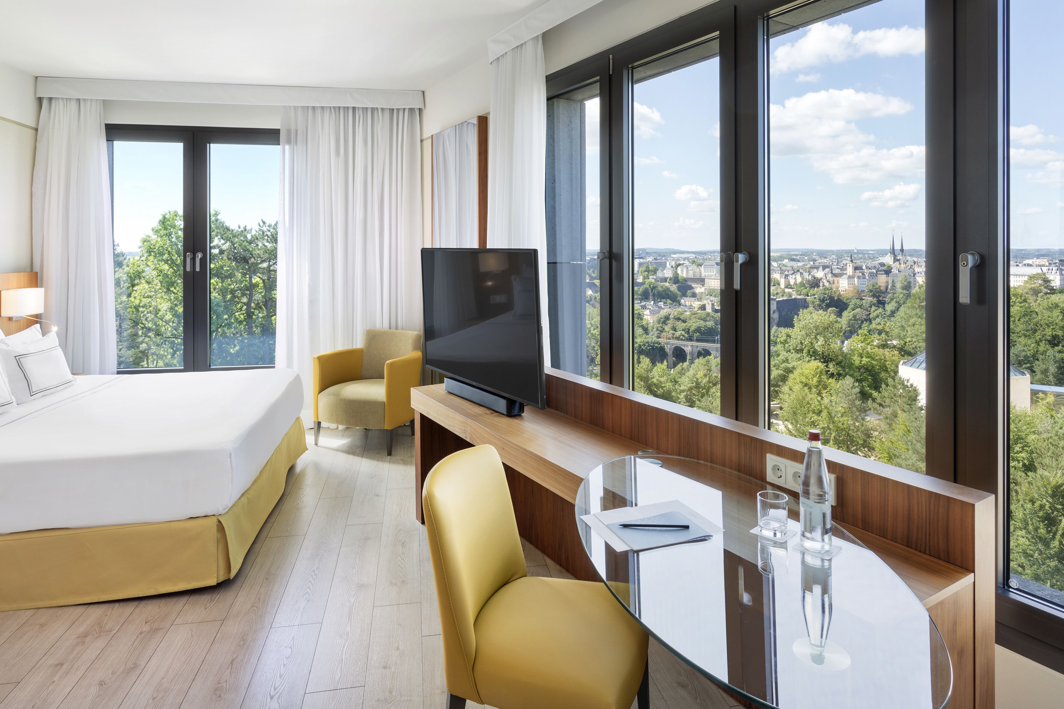 A modern hotel room with a large bed, a TV, a desk with a glass top, and a view of a cityscape through large windows.